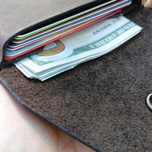 May include: A dark brown leather wallet with a textured interior surface, featuring a metal snap button closure. The wallet contains several stacked credit cards with colorful edges and a folded United States one hundred dollar bill.