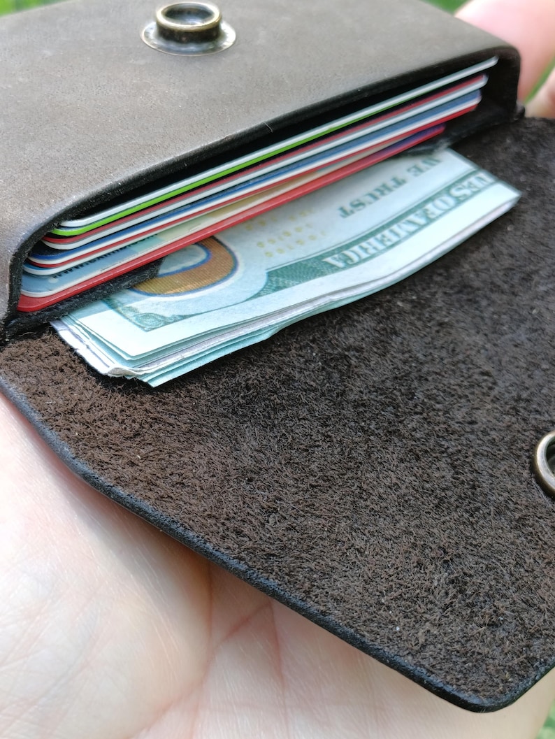 May include: A dark brown leather wallet with a textured interior surface, featuring a metal snap button closure. The wallet contains several stacked credit cards with colorful edges and a folded United States one hundred dollar bill.