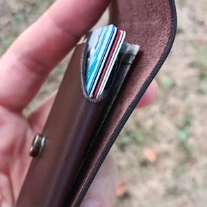 May include: A dark brown leather card holder featuring a flap closure secured with a metallic snap button. The interior compartment contains several stacked credit cards with visible blue, white, and red edges, along with folded paper currency.