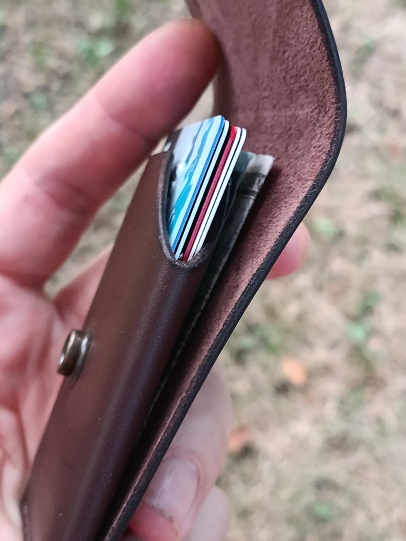 May include: A dark brown leather card holder featuring a flap closure secured with a metallic snap button. The interior compartment contains several stacked credit cards with visible blue, white, and red edges, along with folded paper currency.