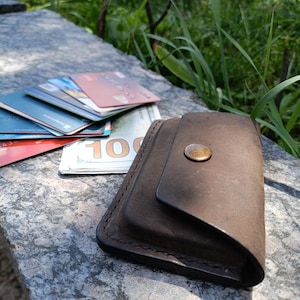 May include: A dark brown leather wallet with a single brass-colored snap button closure. The wallet is positioned next to a stack of various credit cards and a one hundred dollar bill, showcasing a compact design with visible stitched edges.