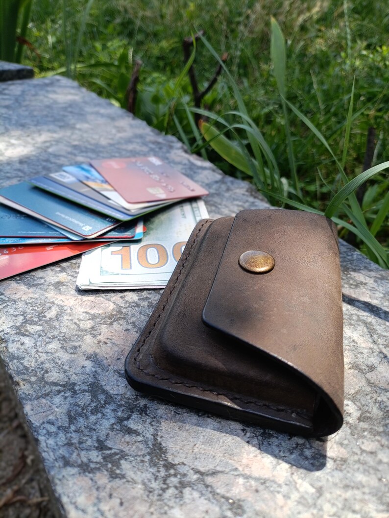 May include: A dark brown leather wallet with a single brass-colored snap button closure. The wallet is positioned next to a stack of various credit cards and a one hundred dollar bill, showcasing a compact design with visible stitched edges.