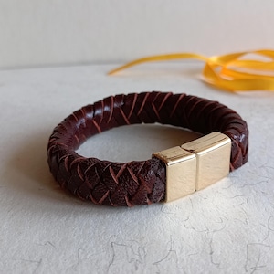 May include: A braided leather bracelet in a rich burgundy color. The bracelet features a gold-colored magnetic clasp. The leather is woven in a tight pattern, creating a textured surface. A stylish accessory.