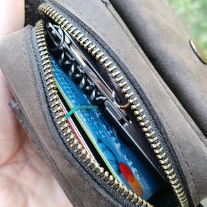 Peut inclure: Une pochette en cuir marron fonc&eacute; avec une fermeture &eacute;clair en m&eacute;tal couleur laiton. Le compartiment ouvert r&eacute;v&egrave;le une carte bancaire bleue avec un logo rouge et jaune, un &eacute;lastique vert fin et un outil multifonction argent&eacute; avec un tire-bouchon noir.