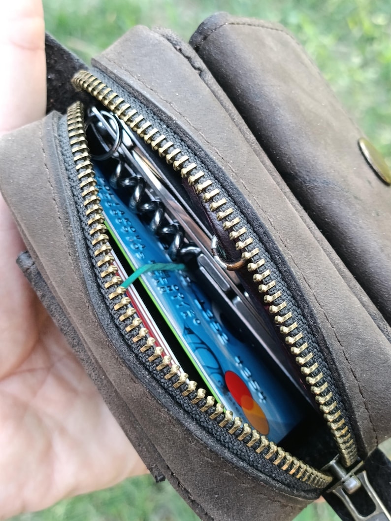 Peut inclure: Une pochette en cuir marron fonc&eacute; avec une fermeture &eacute;clair en m&eacute;tal couleur laiton. Le compartiment ouvert r&eacute;v&egrave;le une carte bancaire bleue avec un logo rouge et jaune, un &eacute;lastique vert fin et un outil multifonction argent&eacute; avec un tire-bouchon noir.