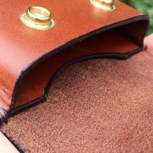 Puede incluir: Una bolsa de cuero color canela con un acabado exterior liso y una superficie interior texturizada. La bolsa se asegura con dos broches de metal dorado en el borde superior, con bordes negros visibles a lo largo de las costuras.