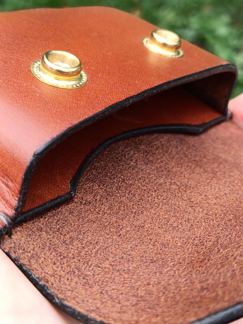 Puede incluir: Una bolsa de cuero color canela con un acabado exterior liso y una superficie interior texturizada. La bolsa se asegura con dos broches de metal dorado en el borde superior, con bordes negros visibles a lo largo de las costuras.