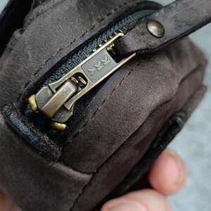 Peut inclure: Une pochette en daim marron fonc&eacute; dot&eacute;e d'une fermeture &eacute;clair YKK couleur laiton. La tirette de la fermeture est constitu&eacute;e d'une bande de cuir marron fonc&eacute; assortie, fix&eacute;e par un petit rivet en laiton. La texture du daim est visible.