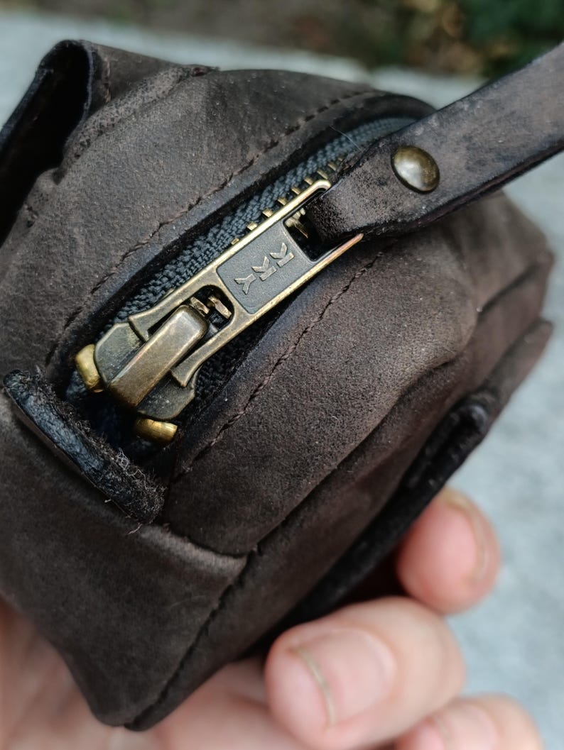 Peut inclure: Une pochette en daim marron fonc&eacute; dot&eacute;e d'une fermeture &eacute;clair YKK couleur laiton. La tirette de la fermeture est constitu&eacute;e d'une bande de cuir marron fonc&eacute; assortie, fix&eacute;e par un petit rivet en laiton. La texture du daim est visible.