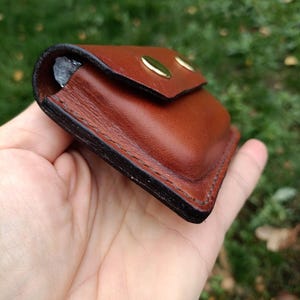 Puede incluir: Una peque&ntilde;a funda rectangular de cuero marr&oacute;n con esquinas redondeadas y bordes oscuros bru&ntilde;idos. La funda cuenta con una solapa asegurada por dos botones de presi&oacute;n de metal dorado y costuras visibles a lo largo del per&iacute;metro.
