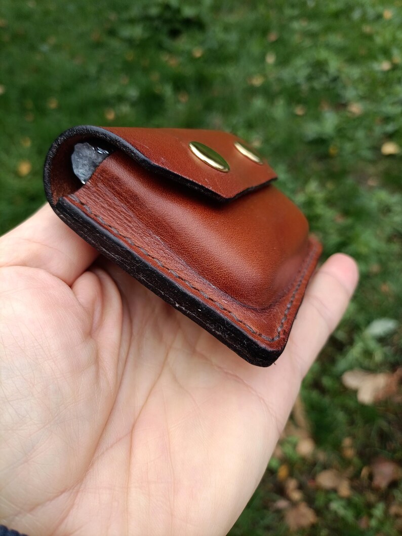 Puede incluir: Una peque&ntilde;a funda rectangular de cuero marr&oacute;n con esquinas redondeadas y bordes oscuros bru&ntilde;idos. La funda cuenta con una solapa asegurada por dos botones de presi&oacute;n de metal dorado y costuras visibles a lo largo del per&iacute;metro.