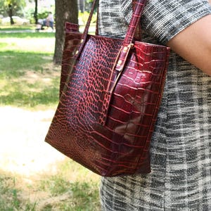 May include: A deep red tote bag with a glossy crocodile skin texture and pattern. The bag features two thin shoulder straps attached with metallic hardware, a rectangular silhouette, and a structured design with a visible sheen on the surface.