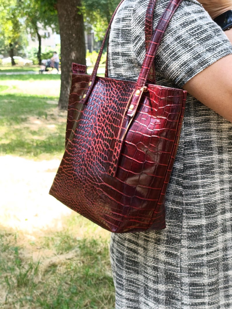 May include: A deep red tote bag with a glossy crocodile skin texture and pattern. The bag features two thin shoulder straps attached with metallic hardware, a rectangular silhouette, and a structured design with a visible sheen on the surface.