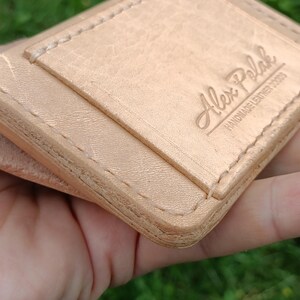 May include: A light tan leather card holder featuring visible stitching along the edges and a debossed logo that reads Alex Polak HANDMADE LEATHER GOODS. The surface of the leather shows a natural grain texture with a smooth, light-colored finish.