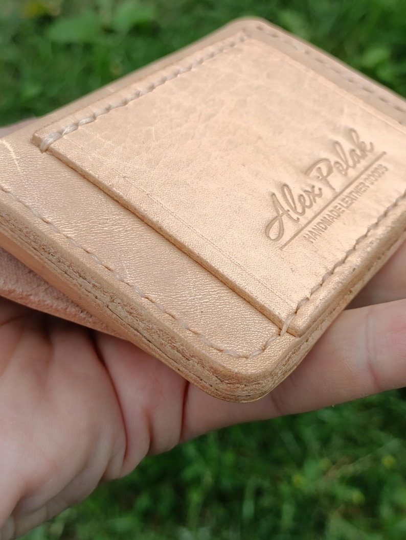 May include: A light tan leather card holder featuring visible stitching along the edges and a debossed logo that reads Alex Polak HANDMADE LEATHER GOODS. The surface of the leather shows a natural grain texture with a smooth, light-colored finish.