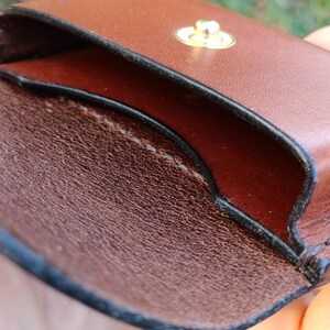 May include: A brown leather pouch with a smooth exterior and a textured interior. The pouch features a gold-toned metal snap button closure at the top centre, with visible stitching along the edges and a dark, finished trim.