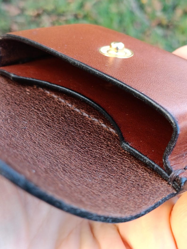 May include: A brown leather pouch with a smooth exterior and a textured interior. The pouch features a gold-toned metal snap button closure at the top centre, with visible stitching along the edges and a dark, finished trim.