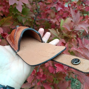 K&ouml;nnte beinhalten: Eine braune Leder-G&uuml;rteltasche mit Druckknopfverschluss. Die Tasche ist offen und zeigt ein hellbraunes Innenfutter. Der Hintergrund zeigt rote und gr&uuml;ne Bl&auml;tter.