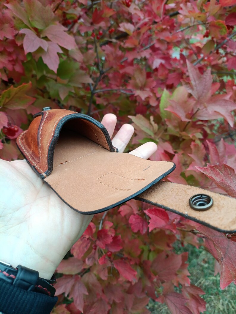 K&ouml;nnte beinhalten: Eine braune Leder-G&uuml;rteltasche mit Druckknopfverschluss. Die Tasche ist offen und zeigt ein hellbraunes Innenfutter. Der Hintergrund zeigt rote und gr&uuml;ne Bl&auml;tter.