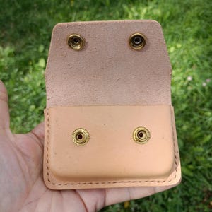 May include: A light tan leather pouch featuring a rectangular shape with rounded corners and visible stitching along the edges. The pouch includes a flap with two brass-colored metal snap fasteners that align with two matching snaps on the body.
