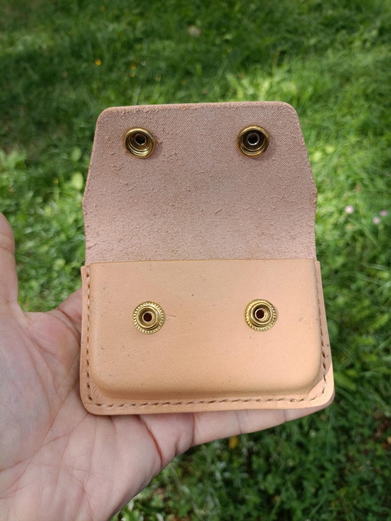 May include: A light tan leather pouch featuring a rectangular shape with rounded corners and visible stitching along the edges. The pouch includes a flap with two brass-colored metal snap fasteners that align with two matching snaps on the body.