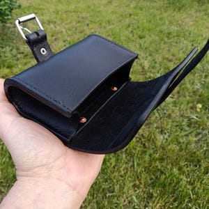 May include: Black leather pouch with a flap closure and a silver-colored buckle. The pouch is open, revealing a dark interior. The strap is visible, and the pouch is held in a hand against a background of green grass.