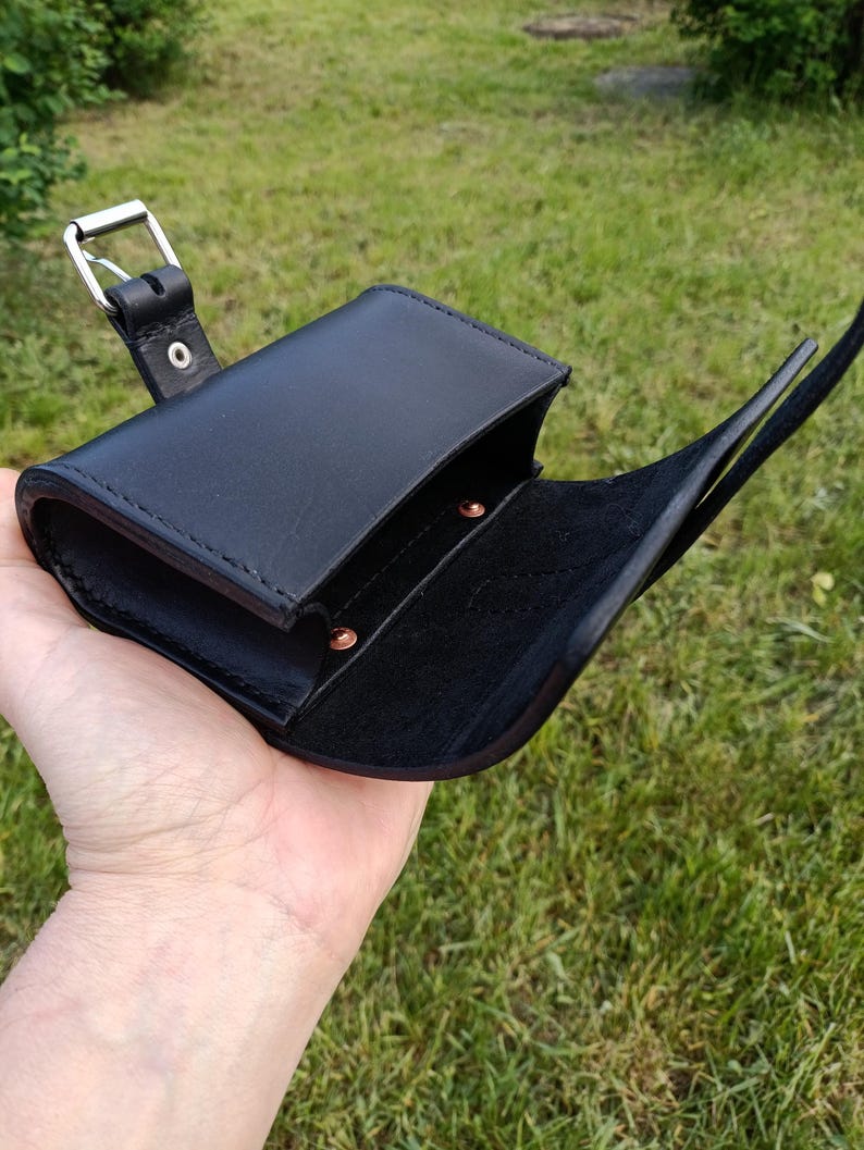 May include: Black leather pouch with a flap closure and a silver-colored buckle. The pouch is open, revealing a dark interior. The strap is visible, and the pouch is held in a hand against a background of green grass.
