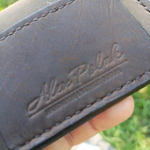 May include: A dark brown leather wallet featuring visible stitching along the edges. The surface is embossed with the text Alex Polak HANDMADE LEATHER GOODS in a script and block font combination, showing a smooth texture and natural grain.