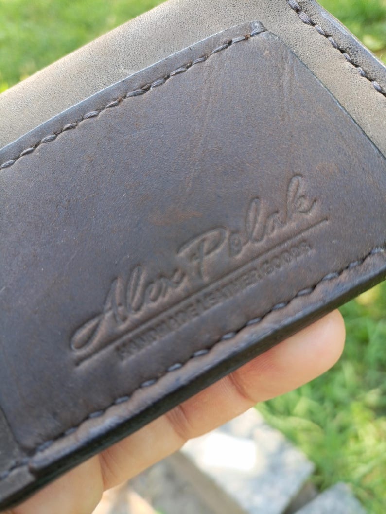 May include: A dark brown leather wallet featuring visible stitching along the edges. The surface is embossed with the text Alex Polak HANDMADE LEATHER GOODS in a script and block font combination, showing a smooth texture and natural grain.