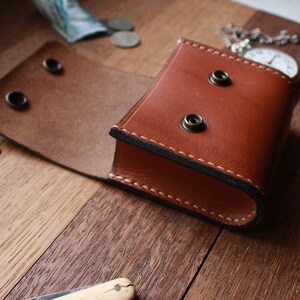 May include: A brown leather pocket watch case with a flap that opens to reveal the inside. The case has two brass rivets on the front and two on the back. The case is sitting on a wooden surface.