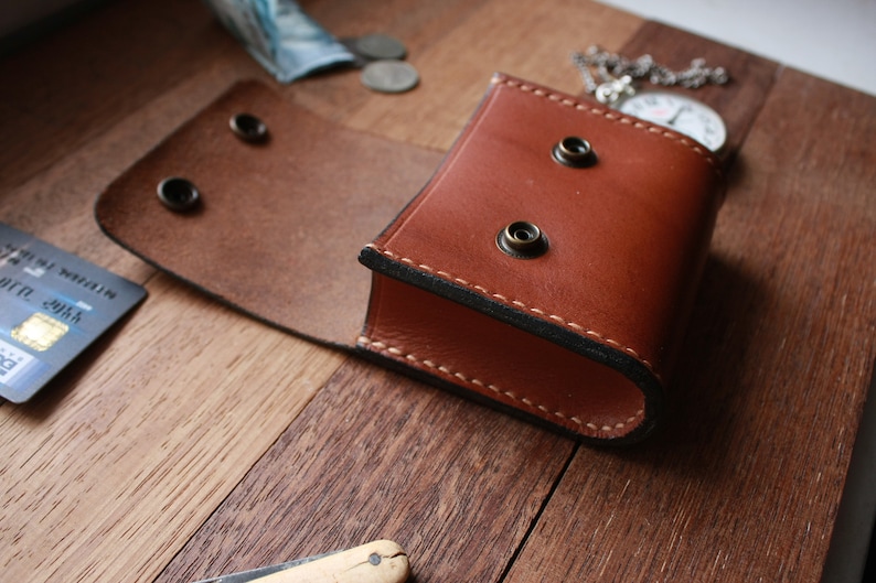 May include: A brown leather pocket watch case with a flap that opens to reveal the inside. The case has two brass rivets on the front and two on the back. The case is sitting on a wooden surface.