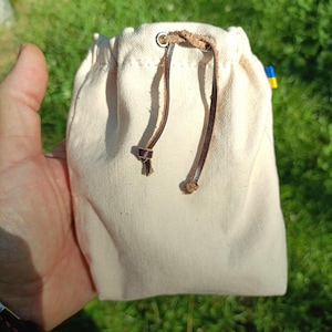 Puede incluir: Una peque&ntilde;a bolsa de lona de color crudo con cierre de cord&oacute;n de cuero marr&oacute;n que pasa a trav&eacute;s de ojales met&aacute;licos. Una diminuta etiqueta de tela azul y amarilla es visible en la costura lateral de la bolsa rectangular de dise&ntilde;o sencillo.