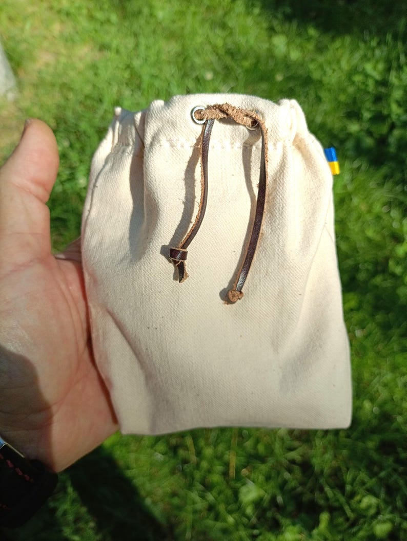 Puede incluir: Una peque&ntilde;a bolsa de lona de color crudo con cierre de cord&oacute;n de cuero marr&oacute;n que pasa a trav&eacute;s de ojales met&aacute;licos. Una diminuta etiqueta de tela azul y amarilla es visible en la costura lateral de la bolsa rectangular de dise&ntilde;o sencillo.