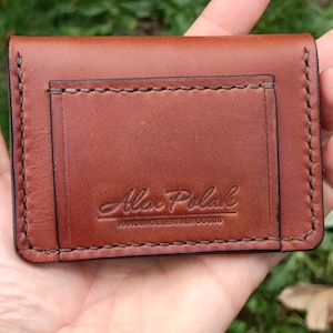 Puede incluir: Un tarjetero rectangular de cuero marr&oacute;n con costuras oscuras visibles a lo largo de los bordes. La parte frontal muestra un logotipo grabado que dice Alex Polak HANDMADE LEATHER GOODS en una combinaci&oacute;n de letra cursiva y de molde.