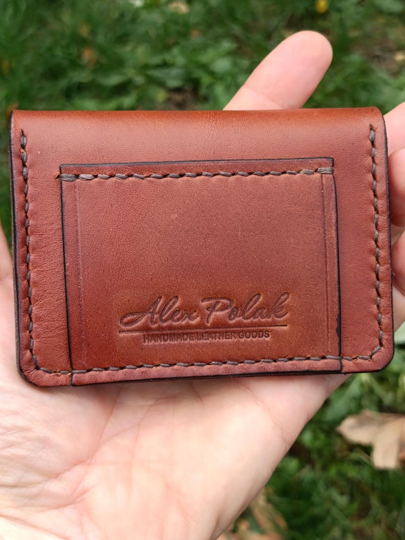 Puede incluir: Un tarjetero rectangular de cuero marr&oacute;n con costuras oscuras visibles a lo largo de los bordes. La parte frontal muestra un logotipo grabado que dice Alex Polak HANDMADE LEATHER GOODS en una combinaci&oacute;n de letra cursiva y de molde.
