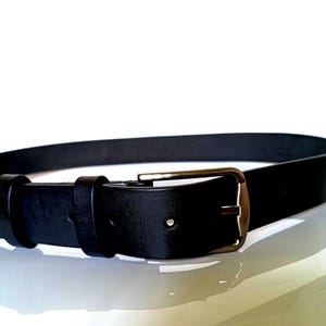 K&ouml;nnte beinhalten: Ein schwarzer Lederg&uuml;rtel mit einer rechteckigen, silberfarbenen Schnalle. Der G&uuml;rtel ist aus glattem, einfarbigem schwarzem Leder gefertigt. Die Schnalle hat ein klassisches Design mit einer einzelnen Dorn.