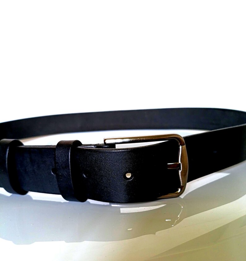 K&ouml;nnte beinhalten: Ein schwarzer Lederg&uuml;rtel mit einer rechteckigen, silberfarbenen Schnalle. Der G&uuml;rtel ist aus glattem, einfarbigem schwarzem Leder gefertigt. Die Schnalle hat ein klassisches Design mit einer einzelnen Dorn.