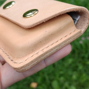 May include: A light tan leather wallet featuring a flap closure secured by two polished brass metal snaps. The wallet is constructed with visible thick stitching along the edges, showcasing the natural texture and layered thickness of the leather.