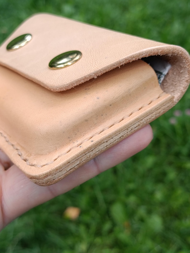 May include: A light tan leather wallet featuring a flap closure secured by two polished brass metal snaps. The wallet is constructed with visible thick stitching along the edges, showcasing the natural texture and layered thickness of the leather.