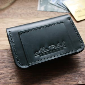 Puede incluir: Una cartera de cuero negro con un borde cosido y el texto "Alex Polak" grabado en la parte delantera.