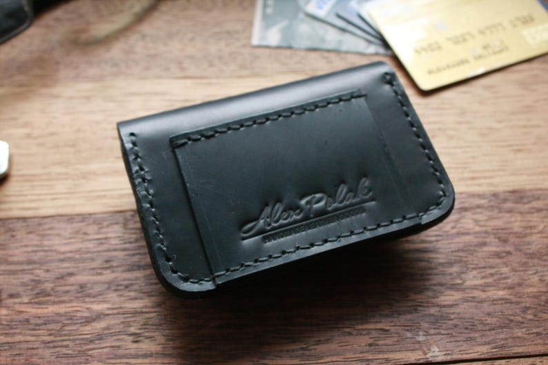 Puede incluir: Una cartera de cuero negro con un borde cosido y el texto "Alex Polak" grabado en la parte delantera.