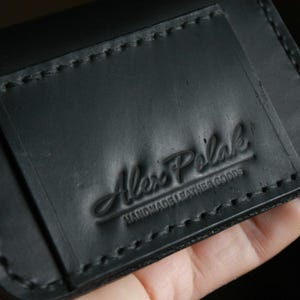 Puede incluir: Cartera de cuero negro con borde cosido y el texto "Alex Polak Handmade Leather Goods" en relieve en la parte delantera.