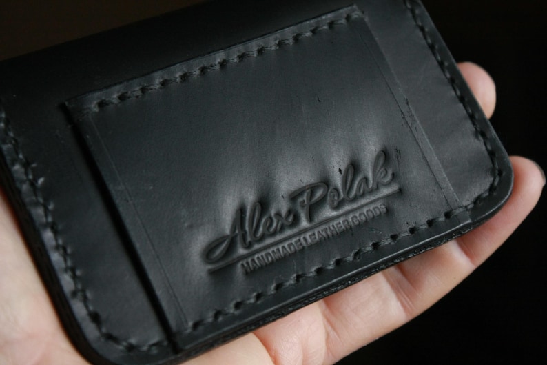 Puede incluir: Cartera de cuero negro con borde cosido y el texto "Alex Polak Handmade Leather Goods" en relieve en la parte delantera.