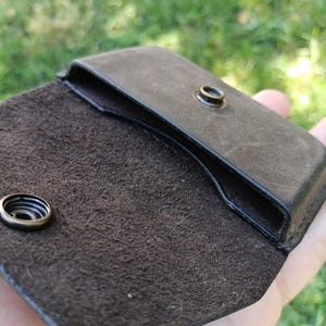 May include: A dark brown leather pouch with a smooth exterior and a textured, suede-like interior. The pouch features a flap closure secured by a circular brass-toned metal snap button, with the corresponding snap component visible on the flap.
