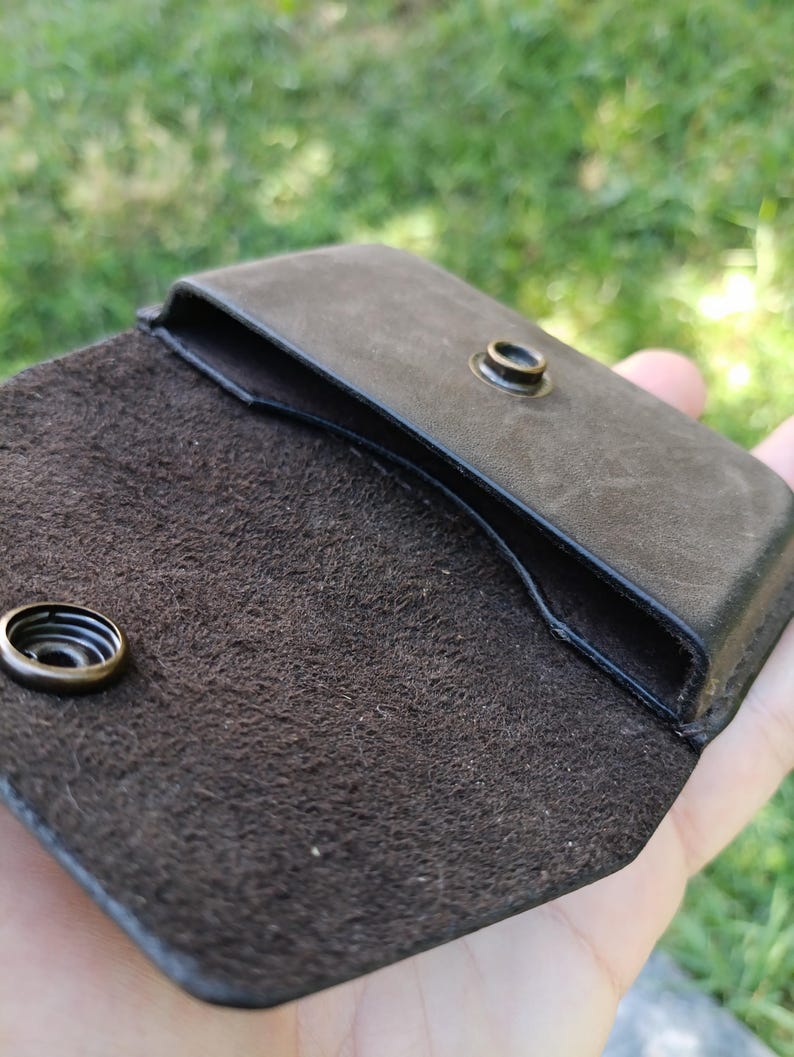 May include: A dark brown leather pouch with a smooth exterior and a textured, suede-like interior. The pouch features a flap closure secured by a circular brass-toned metal snap button, with the corresponding snap component visible on the flap.