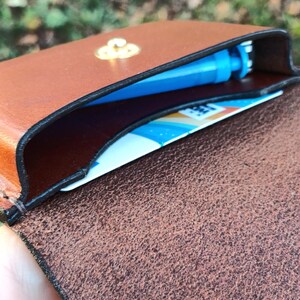 May include: A brown leather card holder with a gold-toned metal snap closure. The interior contains a blue plastic object and a white card with blue and orange graphic elements. The leather has a smooth exterior and a textured interior.