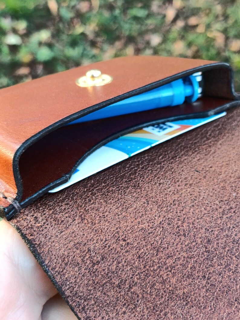 May include: A brown leather card holder with a gold-toned metal snap closure. The interior contains a blue plastic object and a white card with blue and orange graphic elements. The leather has a smooth exterior and a textured interior.