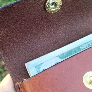 May include: A brown leather wallet featuring a gold-toned metal snap button closure with the text NEW STAR engraved on the fastener. The wallet is partially open, revealing a portion of a paper banknote tucked inside the main compartment.