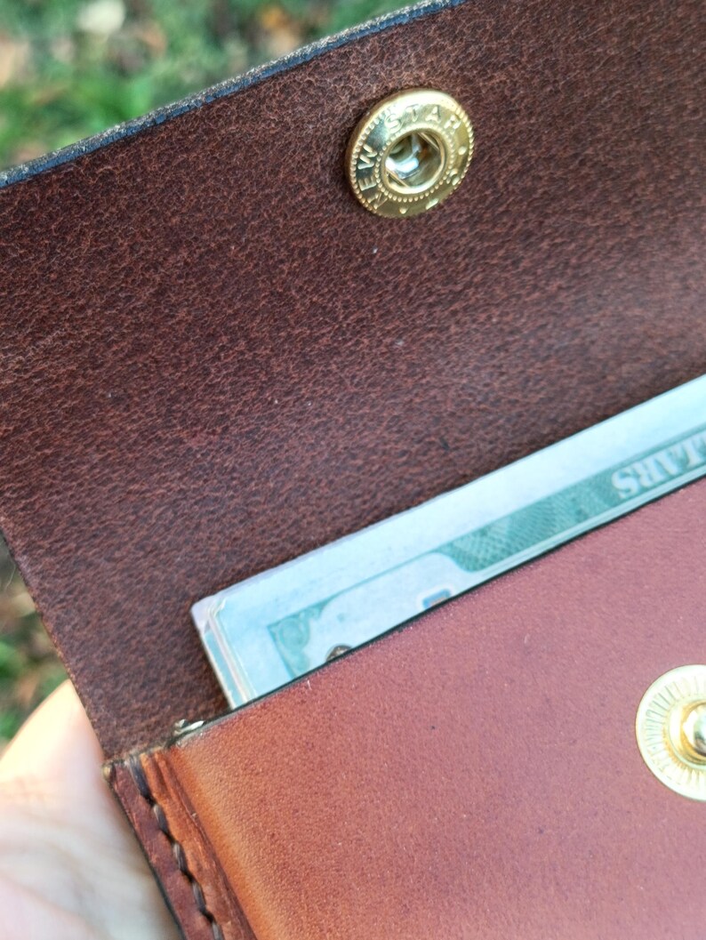May include: A brown leather wallet featuring a gold-toned metal snap button closure with the text NEW STAR engraved on the fastener. The wallet is partially open, revealing a portion of a paper banknote tucked inside the main compartment.