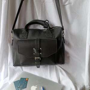 Puede incluir: Un bolso tipo satchel de cuero negro con solapa delantera y cierre de hebilla, asa superior y correa para el hombro desmontable. Tambi&eacute;n se ven un port&aacute;til, tarjetas de cr&eacute;dito y un bol&iacute;grafo.